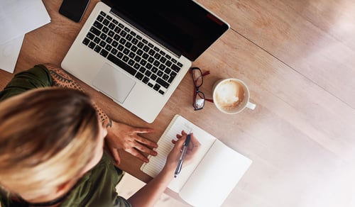 woman at a desk writing a to do list in a notebook with her laptop and coffee shot from above